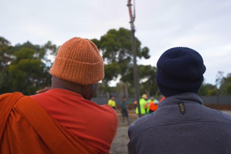 Two men stand looking out onto a construction site where concrete is being poured. One man is in Buddhist religious dress, while the second man is in construction clothing.