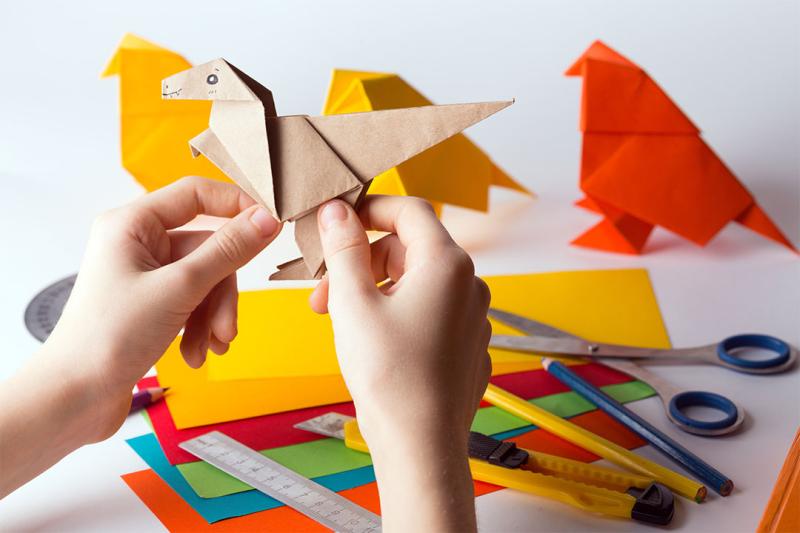 Hand holding an origami dinosaur with other crafting details in the background.