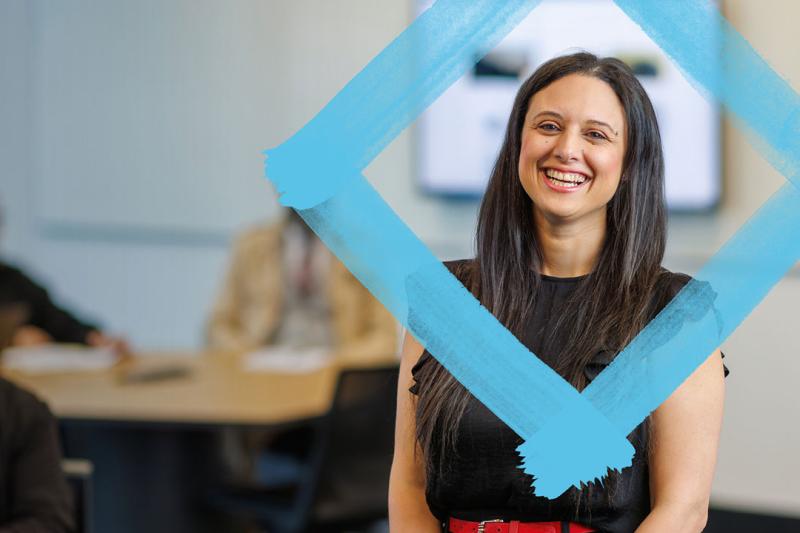 A person standing and smiling at the camera. A blue diamond is painted around their profile. In the background are students sitting around large tables.