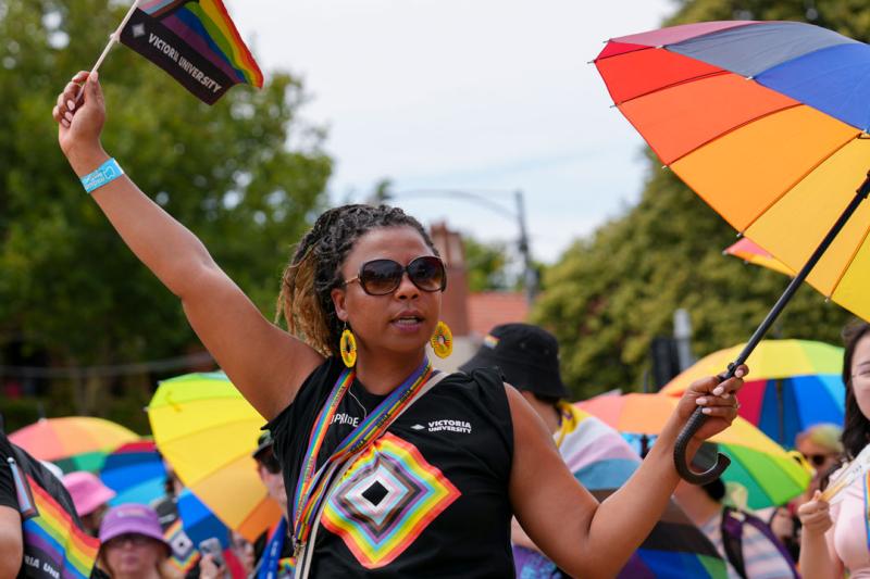 A person holding a pride umbrella and flag outside. They are wearing a pride shirt with the VU logo on it. Other people are standing around them with pride umbrellas.