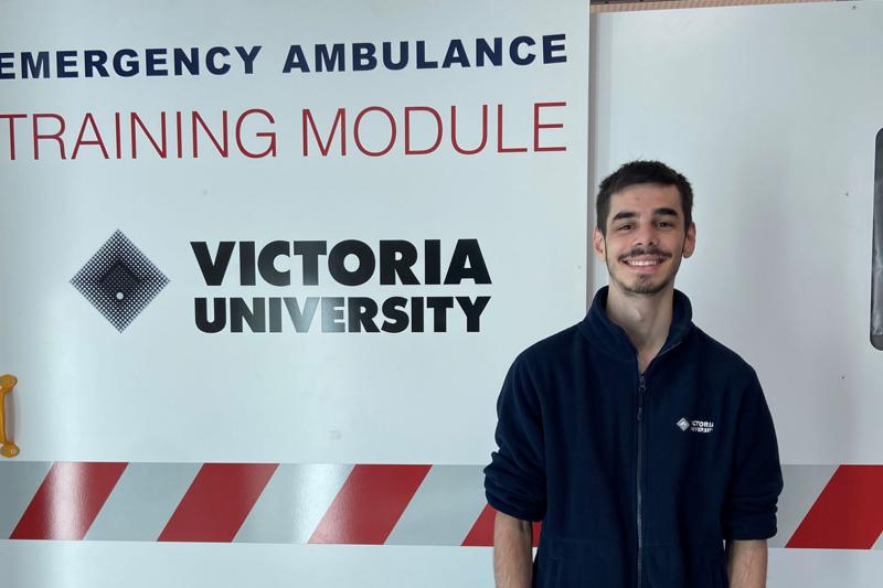 A young man poses by an 'Emergency Ambulance Training Module' bearing a Victoria University logo