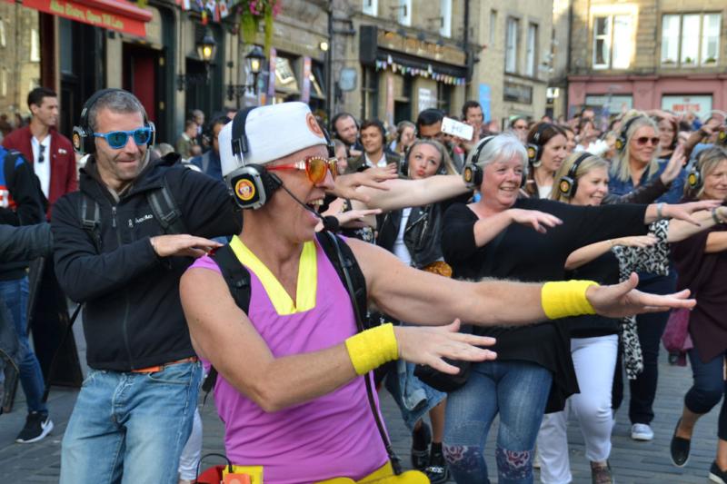 A large group of people outside on the street with headphones on. At the front is a person wearing bright-coloured clothes, glasses and wristguards. They're all coordinating the same dance move.