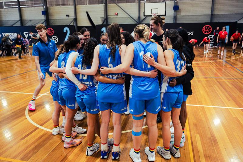 A group of people with long hair in pony tails wearing VU basketball uniforms, rainbow socks and sneakers huddle on a basketball court