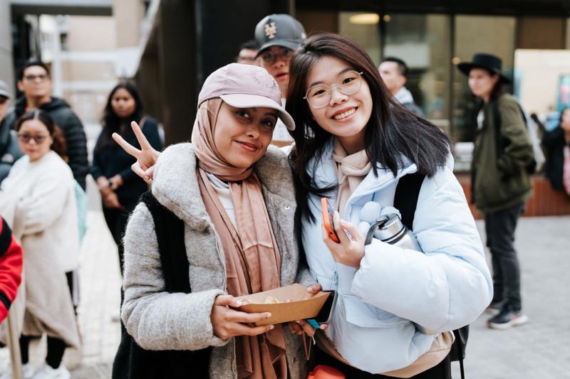 Two students standing outside and smiling at the camera. One student is holding a food carton. Behind them are groups of students walking around.