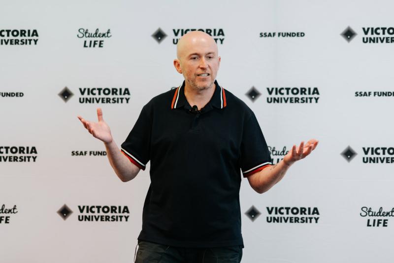 A person speaking at an event. They are standing in front of a wall with Victoria University and Student Life logos.