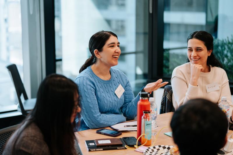 Students at a table. One is responding to a teacher. Another student is looking at the speaking student.