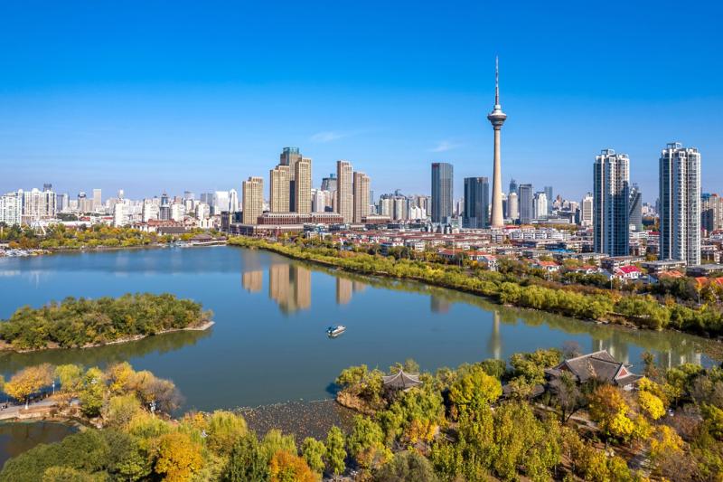 A photo of Shenyang, China. Modern skyscapers and a tower can be seen. Closer to the camera are lush green trees and a small island in a river.
