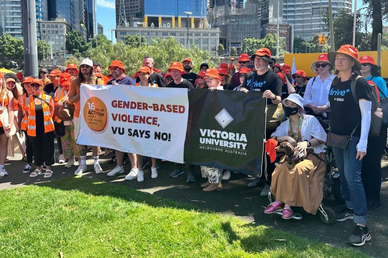 A large group of VU staff and students outside in a park in Melbourne. They're standing behind a large banner that reads "Gender-based violence, VU says No!" with the VU logo and 16 days of activism logo.