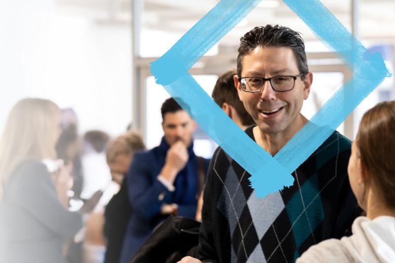 Two people talking in the foreground. A group of event attendees are blurred in the background.