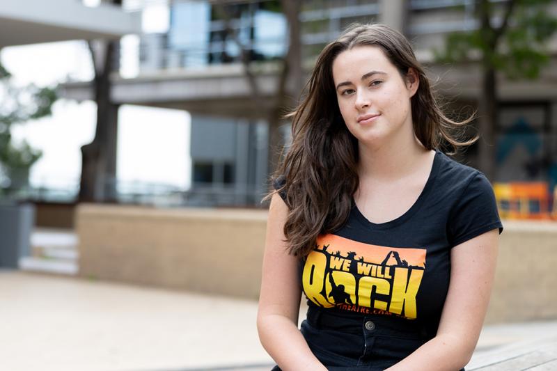 A young student with long hair and a black 'rock' t-shirt sits in a campus courtyard