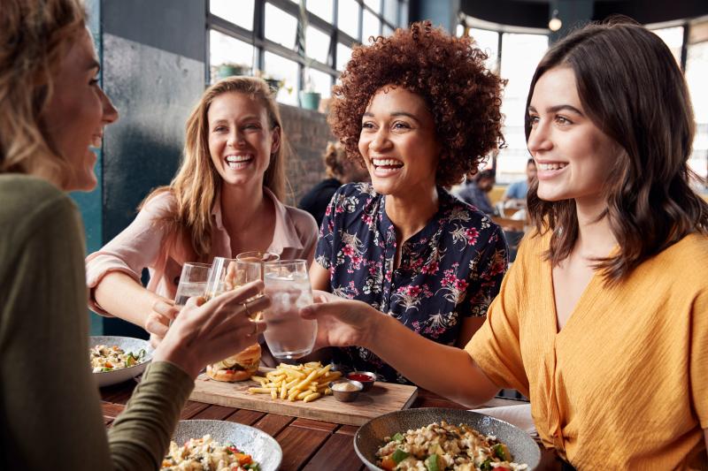 A group of women inside a restaurant smiling and tapping their drink glasses together.