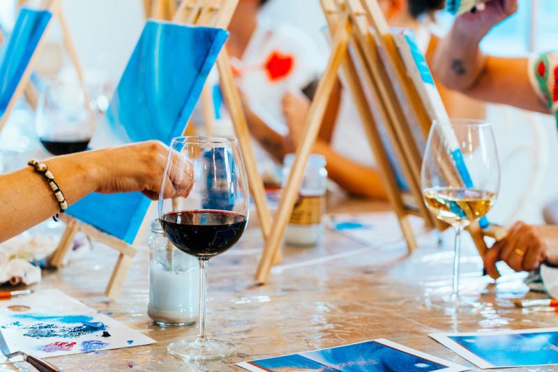A group of people painting on small canvases on a long table. Next to them are glasses of wine.