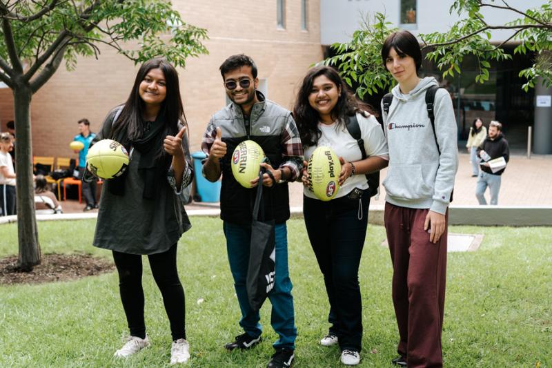Four young adults with backpacks & casual clothes smile and pose in a grassy courtyard