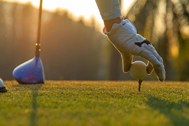 a person puts a golf ball on a tee, on a golf course at sunset.