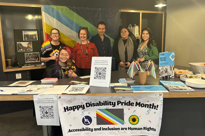 Group of VU staff members gathered around Disability Pride Month stall. There is a large Disabiliiy Pride Flag behind the group. On display in front of the group is a banner with text ' Happy Disability Pride Month' with the wheelchair disability logo, the Sunflower Hidden disability logo and text 'Accessibility and Inclusion are Human Rights'. 