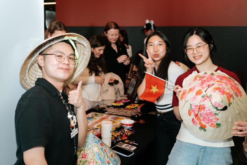 People gathered around a table at an indoor cultural and social event. Participants display decorated conical bamboo hats and a small flag while others socialise in background.