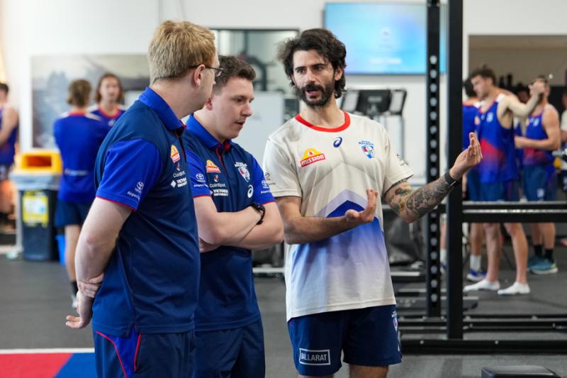 Three people are standing inside a busy gym. One is explaining something and the other two are listening. In the blurred background there are other figures of people in sports clothing and gym equipment.   