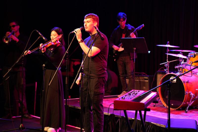 A young musical group with drums, vocal, violin & trumpet perform on a darkened stage with violet lighting