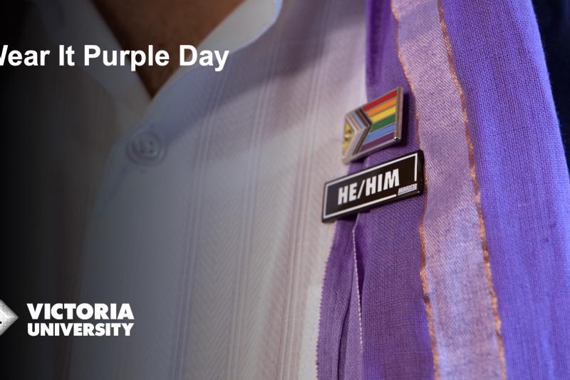 Close up of a person's white shirt and purple scarf, with LGBTQI flag and he/him badges. Text says 'Wear it purple day' and Victoria University logo. 