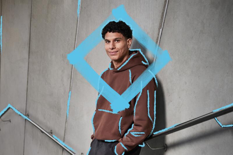 A young man in casual brown hoodie is leaning against a concrete wall of a staircase. He is smiling and relaxed with his hands in his pockets. There is a blue diamond graphic around his head and upper body.   