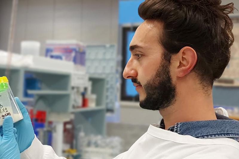 A young male scientist in a white lab coat examines 2 sample containers