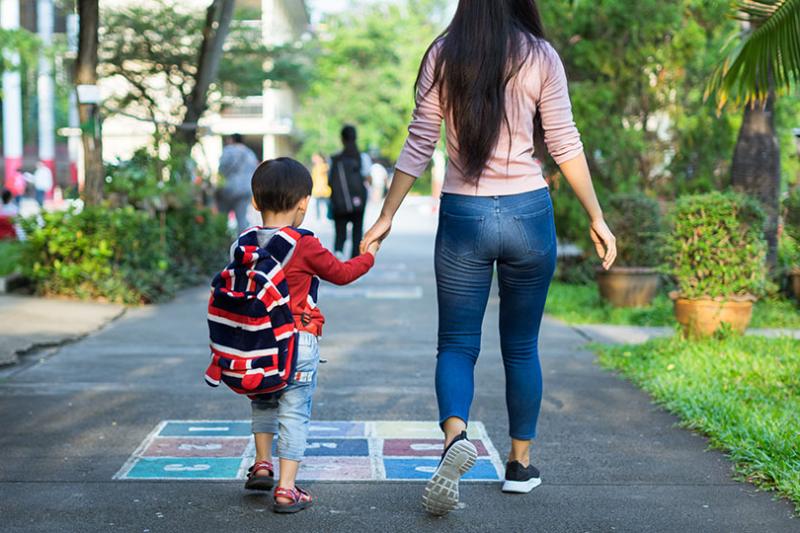 A young boy wearing a backpack walking to school holding his mother's hand.