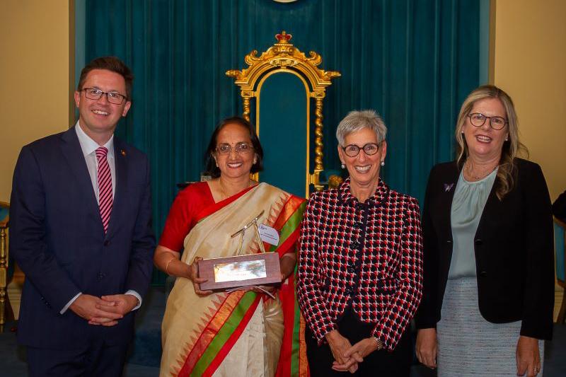 Group of people stand in front of colourful curtain, including woman in traditional Indian dress, holding award