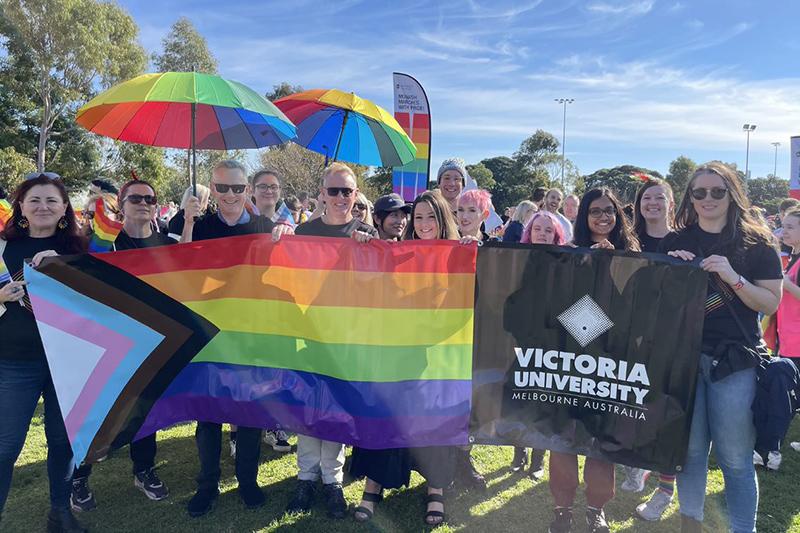 Group of men and women holding rainbow umbrellas, flag with Victoria University logo, at an outdoor gathering