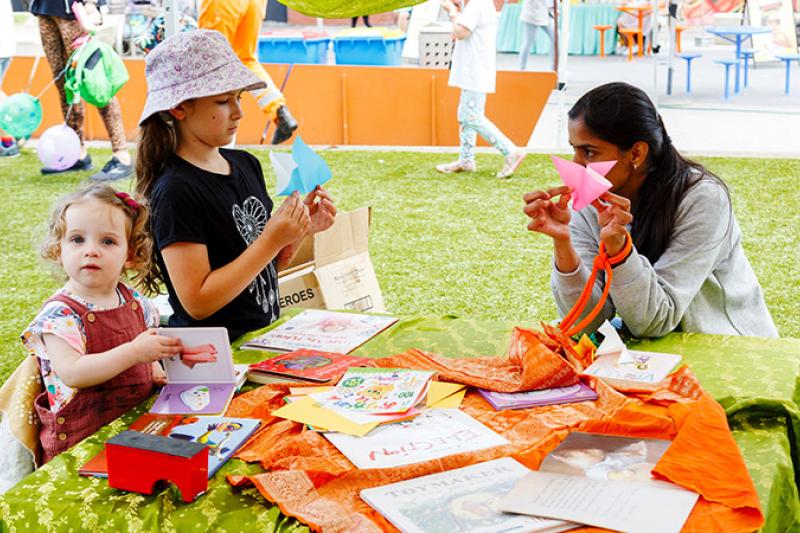 Toddler and child fold paper at an outdoor table, with the help of an adult