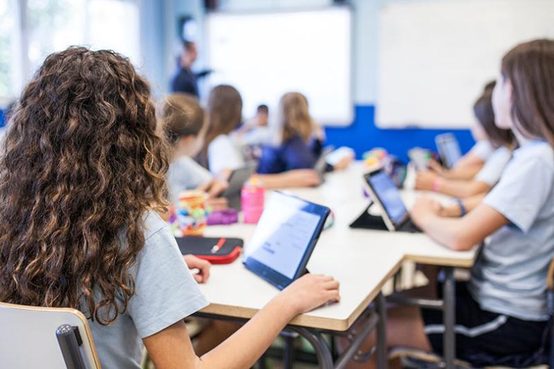  Primary school kids sitting as desks with tablets in a classroom.