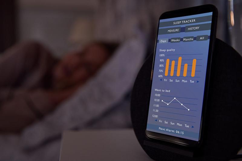 A person sleeps on a bed next to a sleep tracker app on their phone, which is displaying data.
