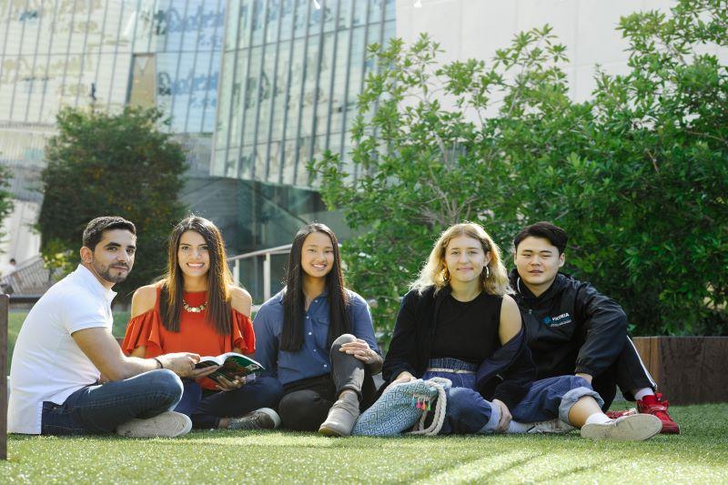 Five diverse, smiling students sitting on the grass at Footscray Park Campus
