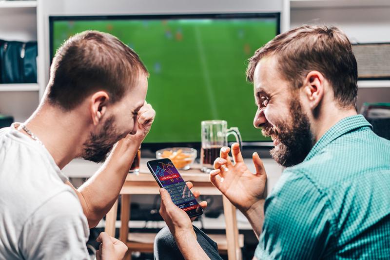  Two friends excitedly checking their sports betting app on their phone, as they watch a soccer match together