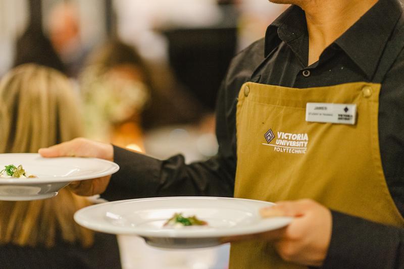 Wait staff in a Victoria University apron carries sophisticated looking dish