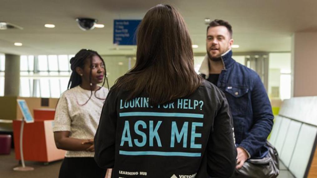 Back of student showing 'Ask me' written on shirt; talking to white male and African-Australian students