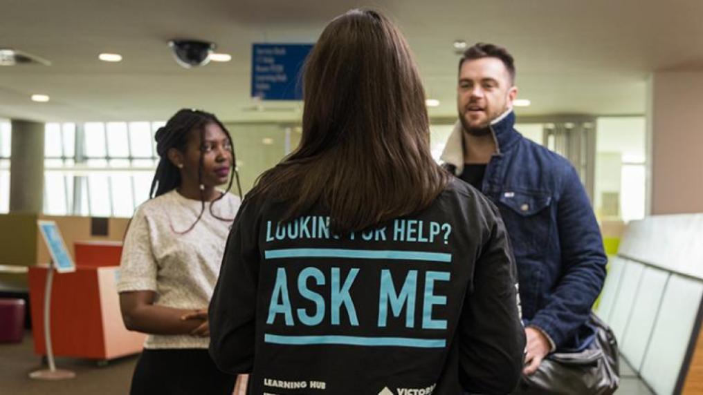 Back of student showing 'Ask me' written on shirt; talking to white male and African-Australian students