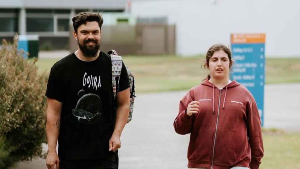 A young man wearing a backpack walks outside with a woman. A Victoria University direction sign is in the background,