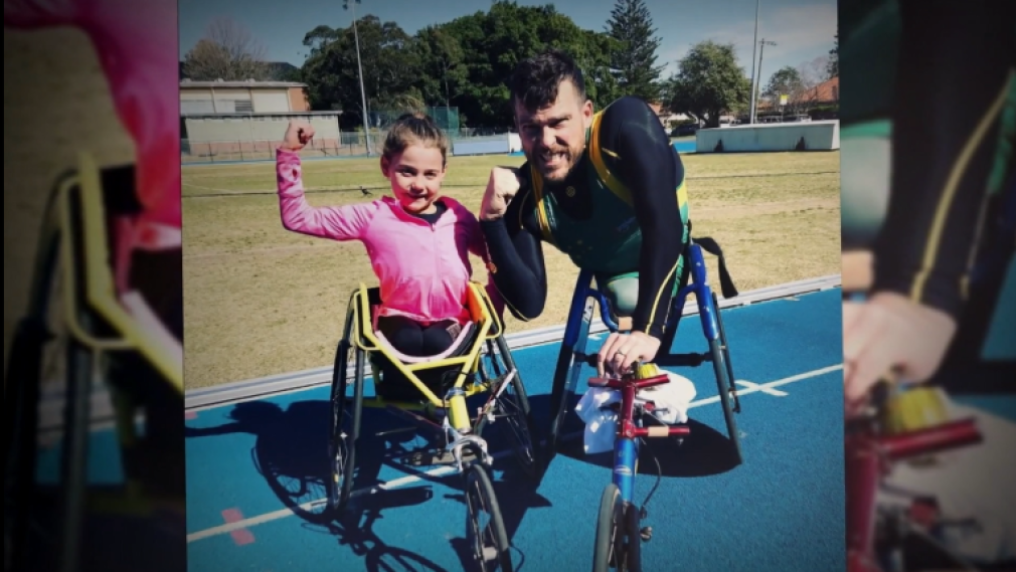 young girl using a 3-wheel racing wheelchair, hand raised in victory, with an adult wheelchair athlete