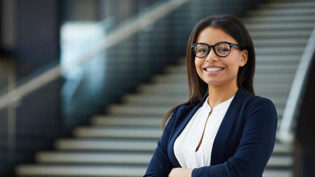 A young woman in glasses standing in a lobby and looking at camera.