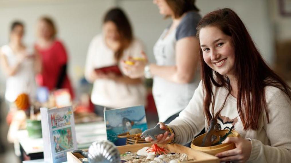 Education student smiles to camera as she sits at table with craft materials, whilst other students mill in the background