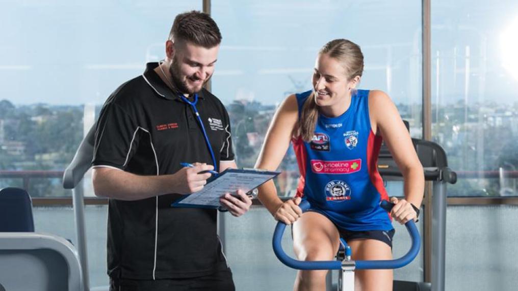 A woman in Western Bulldogs attire on an exercise bike, with a VU staff member standing beside her, with a clipboard and stethoscope