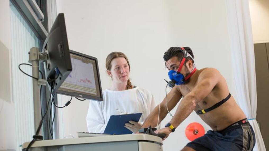 man hooked up to monitoring equipment rides an exercise bike, watched by a woman in a lab coat