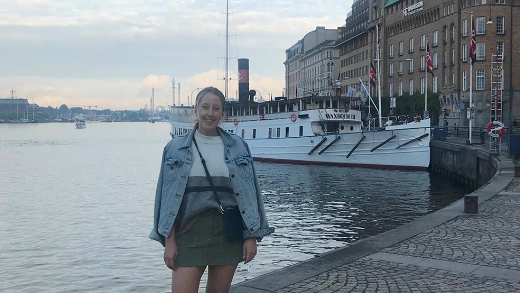Cecilia Pettersson standing and smiling outside, with water, a boat and an old building behind her