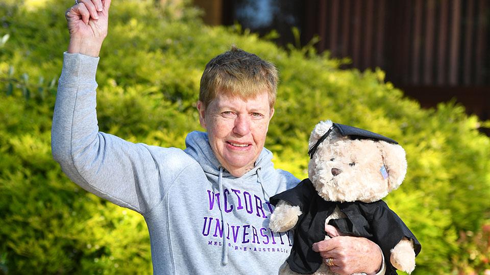 Joan Oliver in a grey VU hoodie, one arm in the air, victorious, holding a VU teddy bear which is dressed in a cap and gown