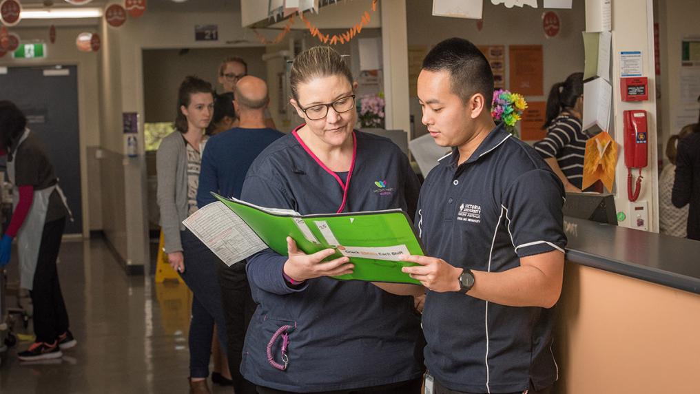 Asian man in Victoria University nursing uniform looks at charts in hospital setting, with woman wearing Western Health Alliance uniform
