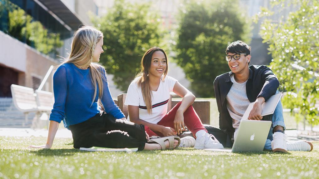 three young student sit smiling on a sunny campus lawn