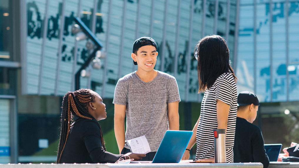 Three happy students, or various ethnicity, smile and talk at a modern campus courtyard