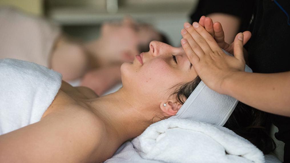 a VU student's hands can be seen massaging the head of a patient who is lying down, eyes closed, covered by a towel with a peaceful look on her face