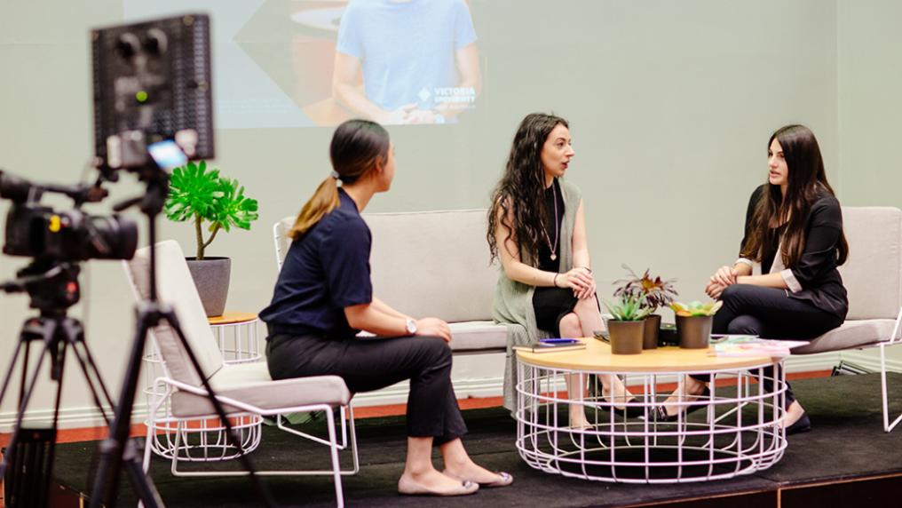 Group of three young adults sit on a film set, in discussion, with screen behind and film camera in the foreground