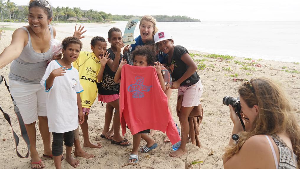 3 young adults pose on a beach with group of Polynesian kids, smiling